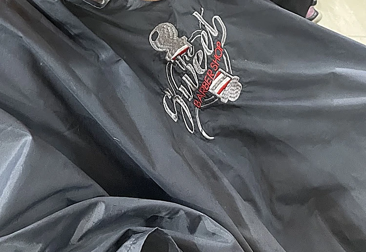 Capa negra de barbero con el logo «Sweet» bordado en blanco y rojo.