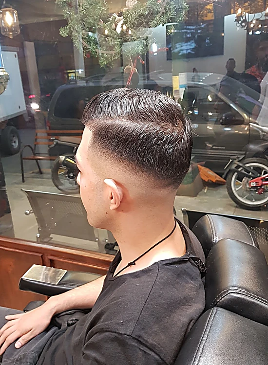 Corte con raya lateral y fade, visto de espaldas en el sillón; al fondo, la ventana a la calle.