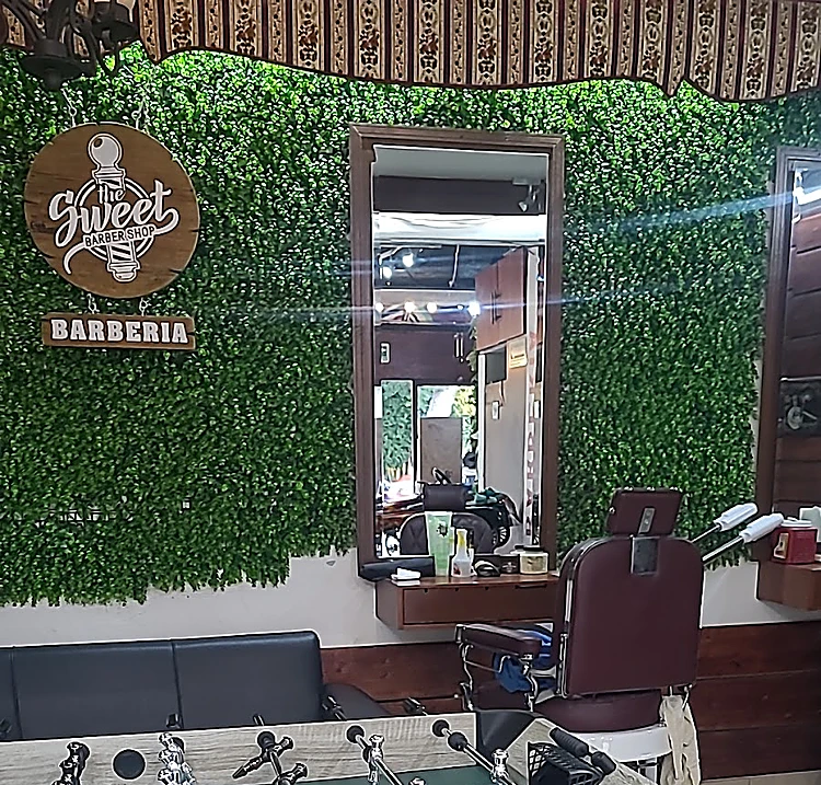 Interior de la barbería: muro de follaje verde con el letrero redondo y la placa «Barbería», un espejo con marco de madera y un sillón de barbero color vino.