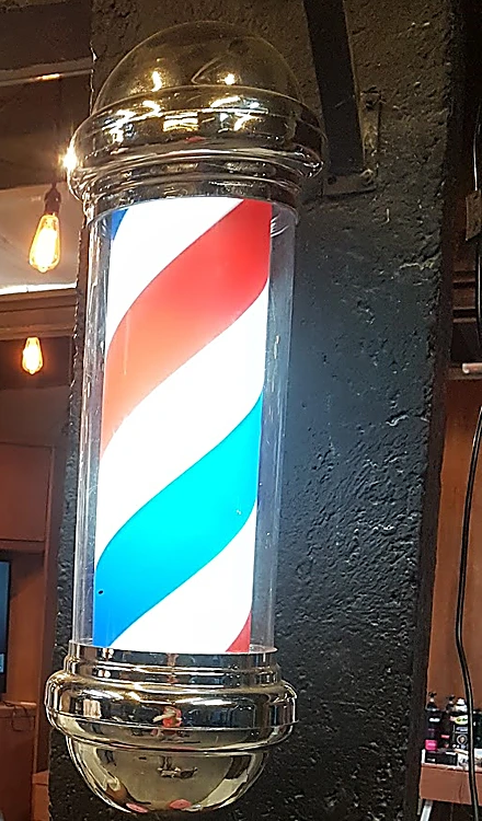 Poste de barbero iluminado, con franjas rojas, blancas y azules y remates cromados, sobre una columna negra.