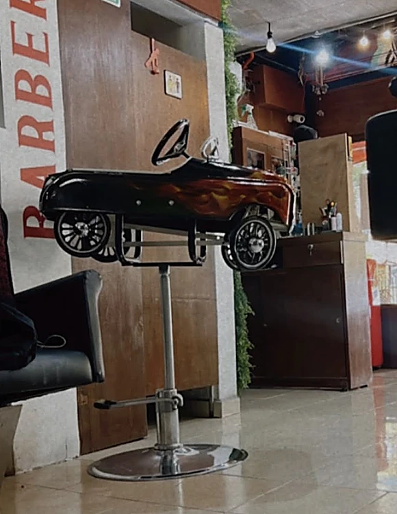 Silla de barbero para niños en forma de carrito negro, montada sobre una base cromada, junto a las letras rojas «Barbería».